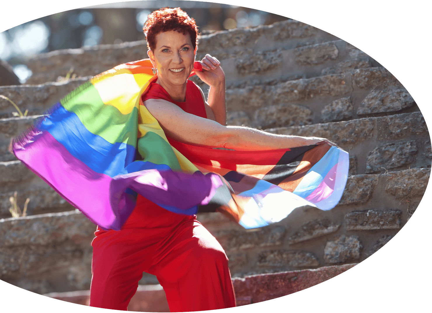 Grace Walcott stands on stone steps outdoors in a red outfit and swings an LGBTQ+ flag