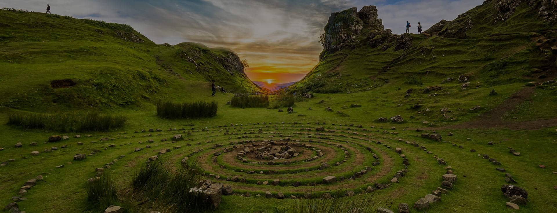 A photo of a spiral labyrinth in the foreground with mountains and sunset in the background. Everything is very green, indicating it might be spring or early summer.