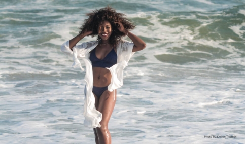 A woman of color is smiling with her arms raised, wearing a black swimsuit stands in the ocean